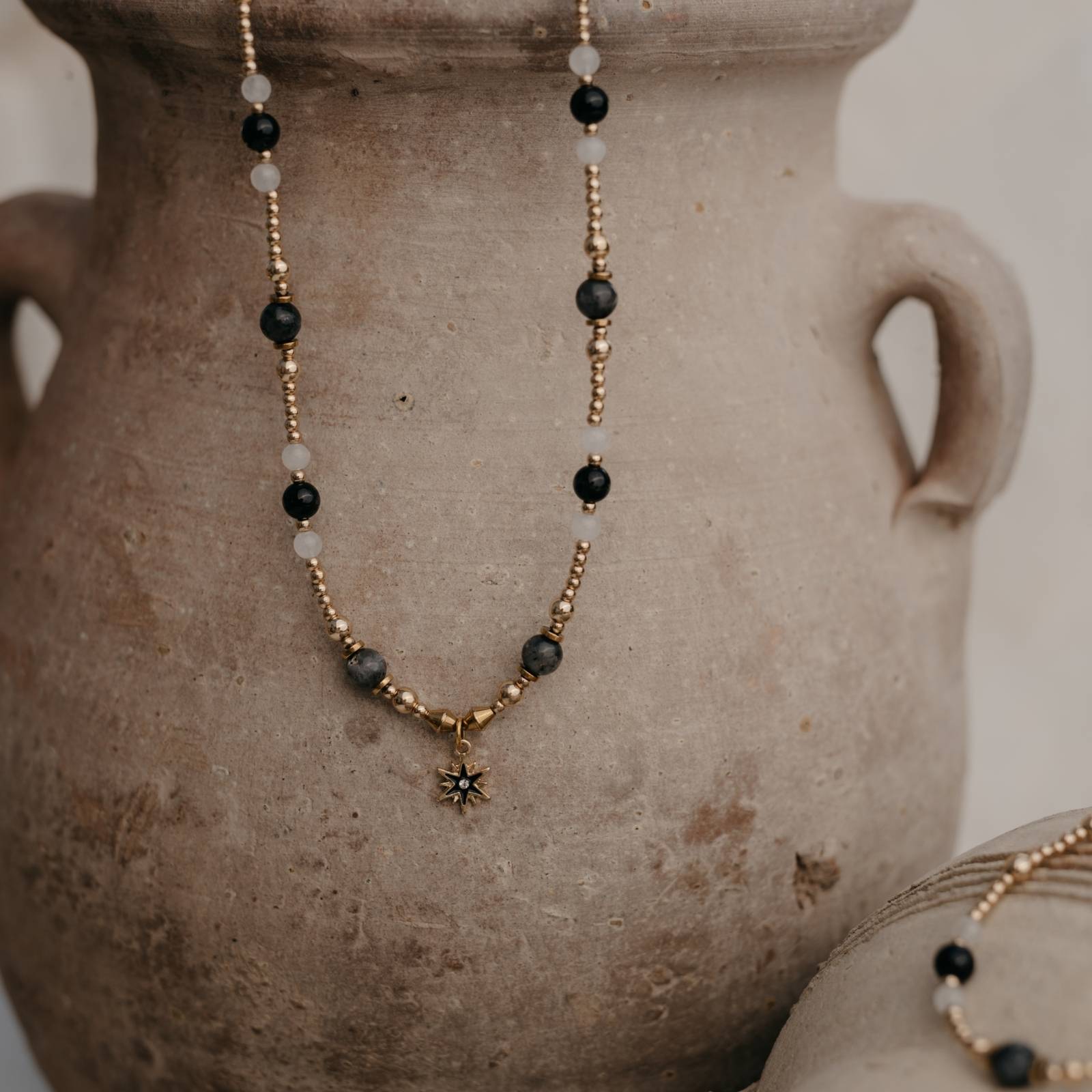 Naszyjnik z labradorytu, hematytu, czarnego agatu i białego jaspisu z zawieszką w kształcie gwiazdy na tle glinianego dzbana — elegancka biżuteria naturalna
