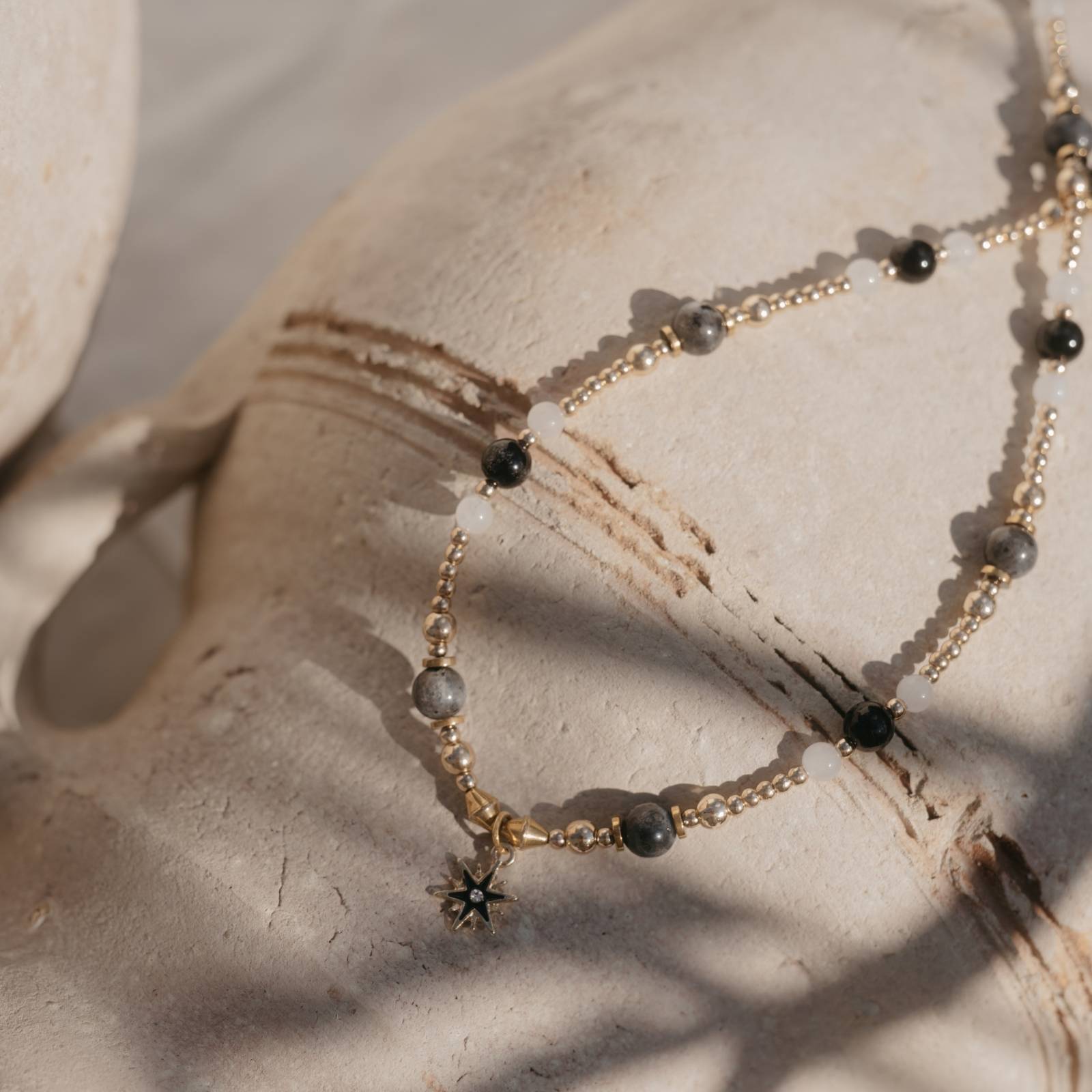 Zbliżenie na kamienie naszyjnika – labradoryt, czarny agat, biały jaspis i złote elementy – w promieniach naturalnego światła na glinianym dzbanie