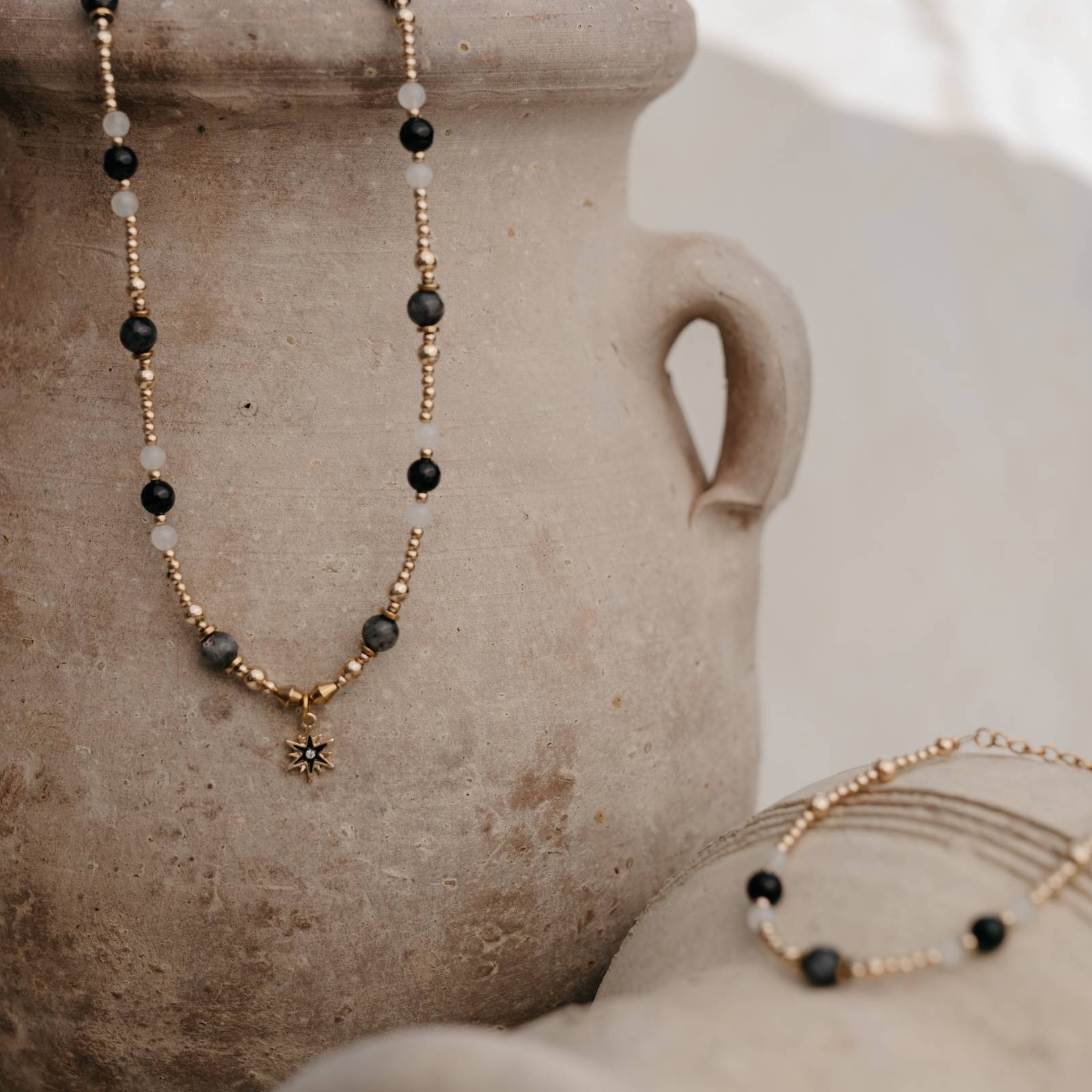 Naszyjnik i bransoletka z kamieni naturalnych w odcieniach złota, czerni i bieli na tle glinianego dzbana – elegancki zestaw biżuterii